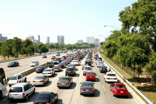 Cars sit in traffic on highway with a city skyline in the background on a sunny day.